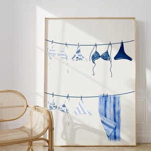 May include: A framed print featuring a watercolor illustration of swimwear hanging on a clothesline. The swimwear includes various blue and white patterned bikini tops and bottoms, and a striped towel. The artwork is displayed in a light-colored wooden frame.