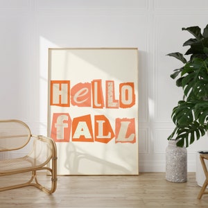 May include: A framed print with the text "hello fall" in orange letters on a white background. The print is hanging on a white wall in a room with a wicker chair and a potted plant.