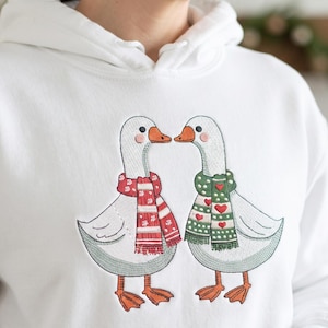 Op de afbeelding: Een wit sweatshirt met capuchon en een geborduurde afbeelding van twee ganzen die naar elkaar toe staan. De ene gans draagt een rood-witte sjaal met patronen, de andere een groen-witte sjaal met rode hartjes. Beide ganzen hebben oranje snavels en poten.