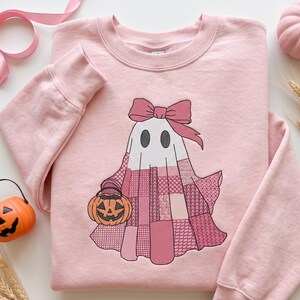 Puede incluir: Una sudadera de cuello redondo rosa claro con un gráfico bordado de un fantasma blanco con un lazo rosa. El cuerpo del fantasma tiene un estilo de retazos en varios tonos de rosa y sostiene un pequeño cubo de dulces de calabaza naranja.