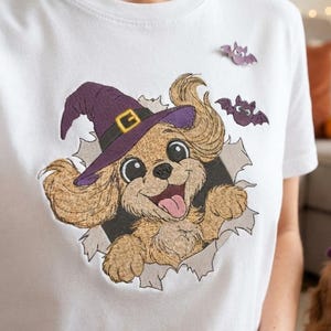 Puede incluir: Una camiseta blanca de manga corta con un gráfico de un perro spaniel de color marrón claro que lleva un sombrero de bruja morado con una hebilla amarilla. El perro emerge de un efecto de papel rasgado, junto a dos pequeños murciélagos morados.
