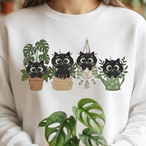 May include: A white long-sleeve sweatshirt featuring a graphic print of four black cats with large green eyes. The cats are positioned inside a terracotta pot, a woven basket, a hanging macrame plant holder, and a green watering can with flowers.