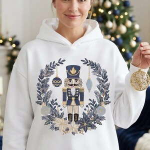 Może przedstawiać: Biała bluza z kapturem z centralną grafiką niebiesko-złotego dziadka do orzechów, otoczonego wieńcem z gałązek sosnowych, gwiazd betlejemskich i wiszących ozdób, z detalami w odcieniach granatu i metalicznego złota.