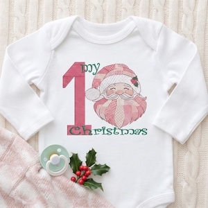 May include: A white long-sleeve baby bodysuit featuring an embroidered design of a pink plaid Santa Claus face next to a large pink number one. The text reads "my 1st Christmas" in green script. A light green pacifier and holly sprigs are visible.