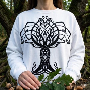 May include: A white long-sleeve sweatshirt featuring a large, black, intricate, symmetrical tree design printed on the front. The graphic consists of flowing, interconnected lines and knotwork patterns that form the shape of a tree with branches.