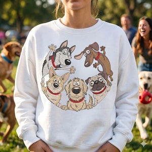 Op de afbeelding: Witte sweatshirt met een cirkelvormig ontwerp van zes cartoonhonden met rode halsbanden. De honden zijn een husky, teckel, corgi, golden retriever en mopshond. De sweatshirt wordt gedragen door een persoon in een parkomgeving.