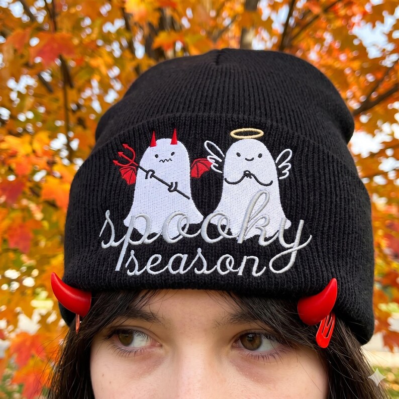 Op de afbeelding: Zwarte gebreide beanie met geborduurde witte geesten, een met duivelshoorns en een drietand, de andere met een halo. De tekst "Spooky Season" is geborduurd in wit script. Rode duivelshoorn haarclips zijn bevestigd.