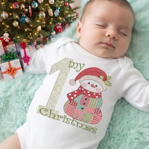 Peut inclure: Un body blanc à manches longues pour bébé avec un grand chiffre un en paillettes dorées et le texte My Christmas. Le motif comprend un bonhomme de neige brodé portant un bonnet et une écharpe rouges avec un motif patchwork rouge, vert et blanc.