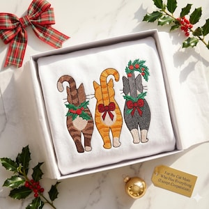 May include: A white sweatshirt with embroidered images of three cats, each adorned with a Christmas wreath and bow. The sweatshirt is presented in a gift box with a plaid bow and holly sprigs. A tag reads: "For the Cat Mom Who Has Everything (Except Cooperation)."