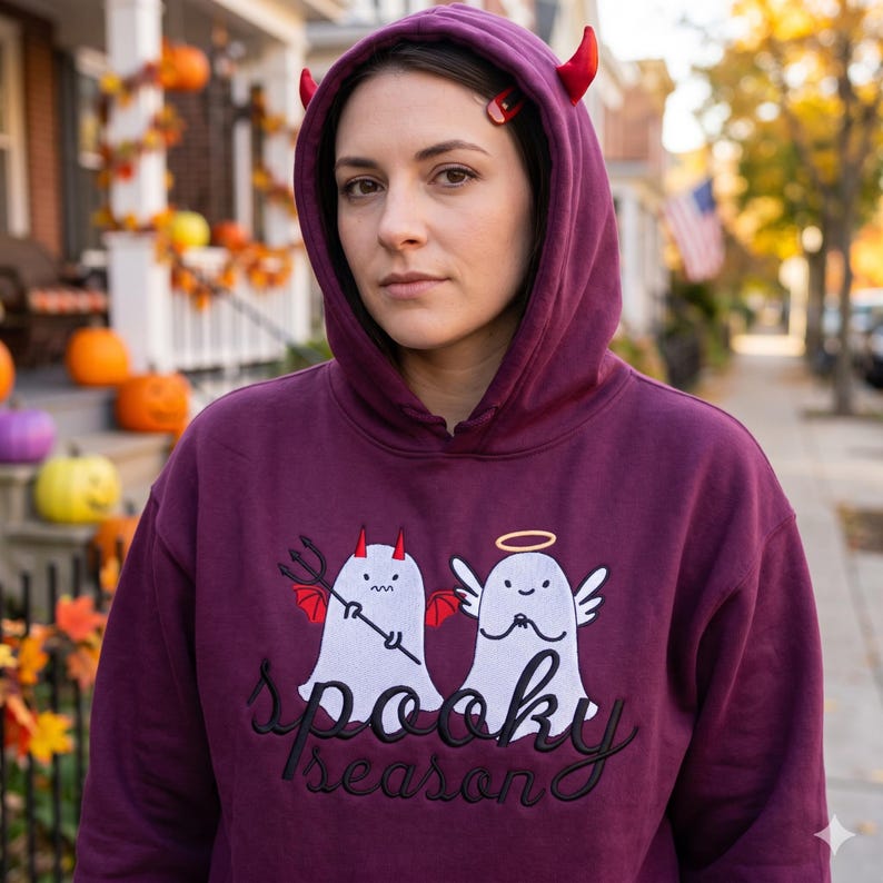 Op de afbeelding: Een bordeauxrode hoodie met capuchon en de woorden "spooky season" in het zwart geborduurd. De hoodie heeft een duivel- en engelgeestontwerp. De persoon op de foto draagt rode duivelshoorns.