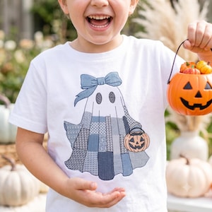 Puede incluir: Una camiseta blanca de manga corta con un gráfico bordado de un fantasma que lleva un lazo azul. El fantasma está diseñado con varios patrones de patchwork en tonos azules y sostiene un pequeño cubo naranja de calabaza para Halloween.