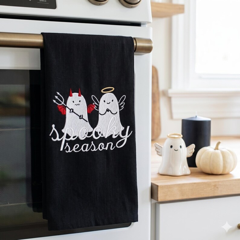 Op de afbeelding: Zwarte keukendoek met geborduurde geesten, een met duivelshoorns en een drietand, de andere met een halo en vleugels. De woorden "Spooky Season" zijn wit geborduurd.