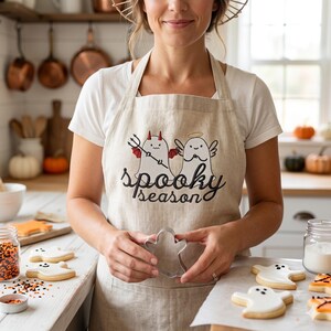 Op de afbeelding: Een beige schort met de tekst "spooky season" en een afbeelding van een spook en een duivel. De schort wordt gedragen door een persoon in een keuken, met Halloween-koekjes en een spookvormige uitsteker.