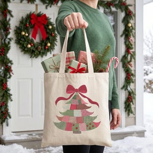 May include: A beige canvas tote bag featuring a printed graphic of a stylized Christmas tree made of various red, green, and white patterned patches, topped with a large red bow. The bag is filled with wrapped gift boxes and pine branches.