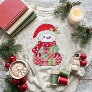May include: A beige crewneck sweatshirt featuring a central embroidered snowman wearing a red Santa hat and a red polka-dot scarf. The snowman body is composed of various green and red patterned quilt-like squares with three small brown buttons.