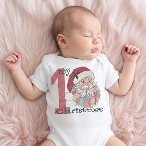May include: A white short-sleeved baby bodysuit featuring an embroidered graphic of a Santa Claus face with a red hat and the text "my 1st Christmas" in red and green thread, with the number one styled in a textured red pattern.