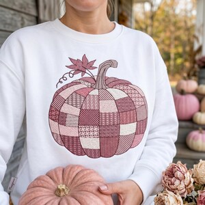 May include: White sweatshirt featuring a patchwork pumpkin design in shades of pink and burgundy. The pumpkin has a stem, leaf, and tendrils. The person is holding a pink pumpkin. The sweatshirt is a fall-themed item.