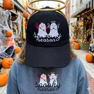 Op de afbeelding: Zwarte baseballcap met de geborduurde tekst "Spooky Season" en een cartoon duivel- en engelgeestontwerp. Een gouden halo staat boven de pet. Hetzelfde ontwerp staat op een blauwe sweatshirt.