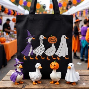 May include: Black tote bag featuring embroidered Halloween geese in witch, mummy, jack-o'-lantern, and ghost costumes. Matching goose figurines in the same costumes are displayed in front of the bag. The scene is set at an outdoor market.