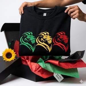 May include: A black crewneck sweatshirt featuring three embroidered lion head profiles in green, yellow, and red. The sweatshirt is presented in a black gift box with green and red tissue paper, a yellow sunflower, and a tag reading June 19th. Always.