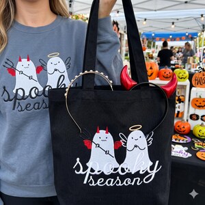 Op de afbeelding: Zwarte draagtas met een wit geborduurd ontwerp van twee geesten, een met duivelshoorns en de andere met een halo, en de woorden "Spooky Season". De tas bevat een gouden haarband en rode duivelshoorns.