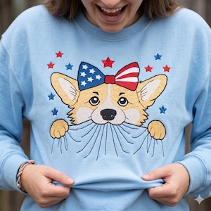 May include: A light blue crewneck sweatshirt featuring an embroidered design of a tan and white Corgi dog wearing a red, white, and blue star-and-stripe patterned bow. The dog is depicted pulling at the fabric, surrounded by small red and blue stars.