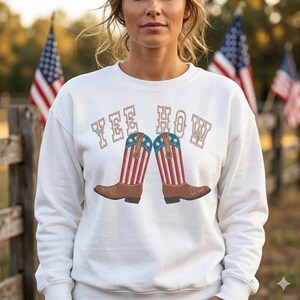 May include: White sweatshirt with the text "YEE HOW" in a Western-style font. The design features a pair of cowboy boots with red and white stripes and blue stars, evoking a patriotic theme. American flags are visible in the background.