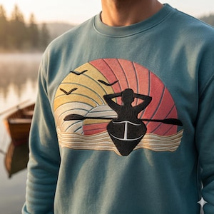 Puede incluir: Sudadera de color turquesa con un diseño bordado de una persona en un bote con remos, sobre un fondo de puesta de sol con pájaros. El diseño incorpora tonos de naranja, amarillo y negro. La sudadera es de material suave.