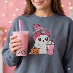May include: A slate blue crewneck sweatshirt featuring an embroidered graphic of a white ghost wearing a pink and grey striped beanie with the text BOO. The ghost holds a small orange jack-o'-lantern and a cup of pink bubble tea with a straw.