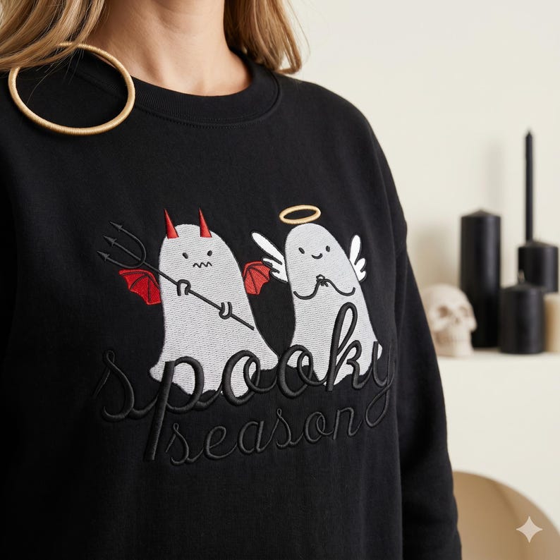 Op de afbeelding: Zwart sweatshirt met geborduurd ontwerp met twee cartoongeesten, een als duivel met een drietand en de andere als engel met een halo. De tekst "Spooky Season" is eronder geborduurd. Een goudkleurig cirkelvormig object is zichtbaar.