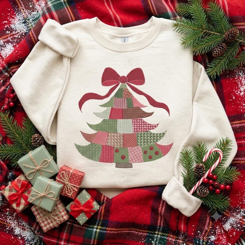 May include: A cream-colored crewneck sweatshirt featuring a graphic of a Christmas tree made of various patchwork patterns in shades of green, red, and pink, topped with a large red bow. The design is centered on the front of the garment.