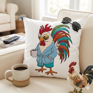 Puede incluir: Cojín cuadrado blanco con un diseño de gallo bordado. El gallo lleva una bata azul y sostiene una taza de café. Gafas de sol negras descansan sobre el cojín. También se ve una figura de gallo de cerámica y una taza de café.