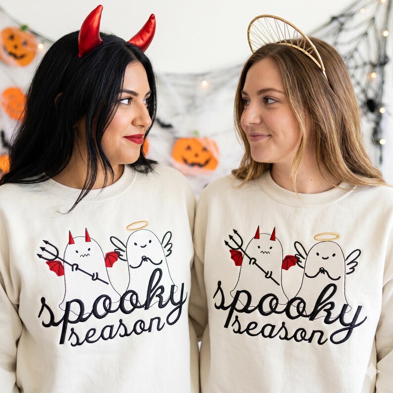 Op de afbeelding: Twee vrouwen in bijpassende cr&egrave;mekleurige sweatshirts met een "spooky season"-afbeelding. De afbeelding toont een duivel en een engel als geesten. E&eacute;n vrouw draagt rode duivelshoorns, de ander een gouden halo-hoofdband. Halloween-thema.