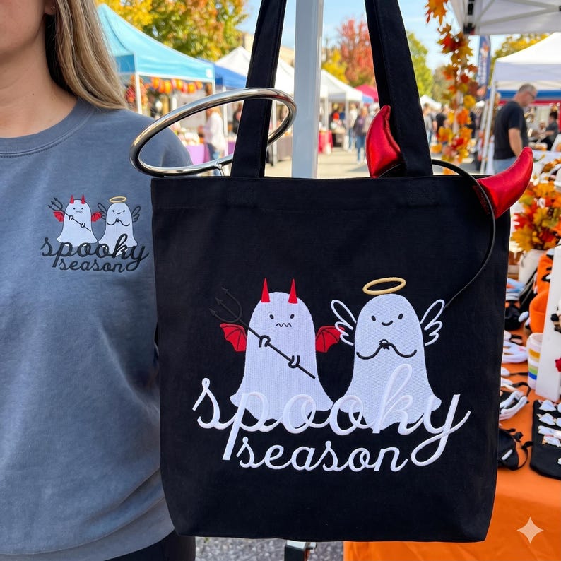 Op de afbeelding: Zwarte draagtas met witte geborduurde spookfiguren, een met duivelshoorns en een drietand, de andere met een halo en vleugels. De tekst "Spooky Season" is eronder geborduurd. Een grijze sweatshirt met hetzelfde ontwerp is zichtbaar.