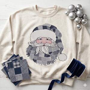 May include: Cream-colored sweatshirt featuring an embroidered Santa Claus face with a patchwork hat and beard. The Santa design incorporates shades of gray and blue. Decorative silver ornaments and blue velvet ribbon are also present.