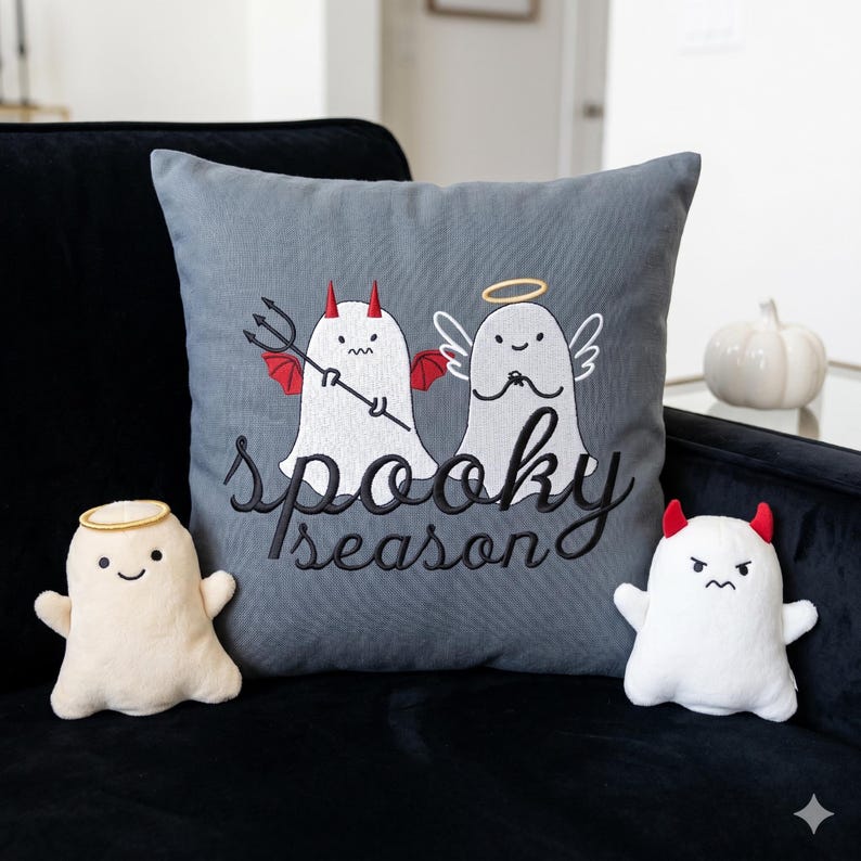 Op de afbeelding: Een grijs decoratief kussen met geborduurde geesten, een met een halo en vleugels, de andere met hoorns en een drietand. Op het kussen staat "spooky season" in zwart script. Er zijn ook twee pluche spookfiguren aanwezig.
