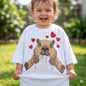 Könnte beinhalten: Weißes T-Shirt mit einem braunen Capybara-Design und roten Herzen. Das Capybara ist mit einem gelangweilten Gesichtsausdruck gestickt und zieht am Shirt. Das Shirt wird von einem Kind getragen.