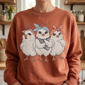 May include: Rust-coloured sweatshirt with an embroidered design of three cartoon chickens. Each chicken wears a bandana and a bow. The chickens have white feathers, brown eyes, and orange beaks and feet. The sweatshirt has a crew neck and long sleeves.