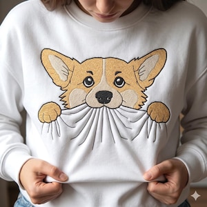 May include: White sweatshirt with an embroidered corgi dog design. The corgi is light brown and white, with large ears and a black nose. The dog appears to be holding the sweatshirt material in its mouth.