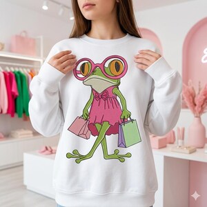 Könnte beinhalten: Weißes Sweatshirt mit einem Cartoon-Frosch-Design. Der grüne Frosch trägt eine rosa Brille und ein rosa Kleid und trägt Einkaufstaschen. Das Sweatshirt hat lange Ärmel.