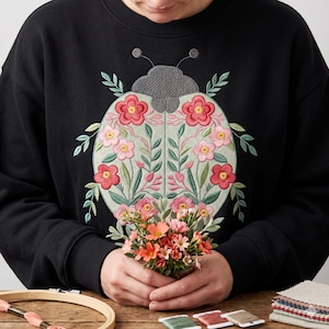 May include: Black sweatshirt with an embroidered ladybug design. The ladybug has a gray head and a light green body adorned with pink and red flowers and green leaves. The person is holding a small bouquet of orange and pink flowers.