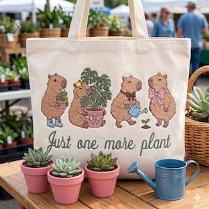 Könnte beinhalten: Cremefarbene Tragetasche mit einer Illustration von Capybaras und dem Text "Just one more plant". Das Bild zeigt drei kleine rosa Blumentöpfe mit Sukkulenten und eine blaue Gießkanne. Die Tasche hat cremefarbene Griffe.