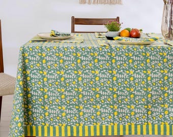 May include: A green tablecloth featuring a pattern of yellow lemons and grey leaves. The tablecloth has a yellow and green striped border. The table is set with matching placemats, napkins, and plates. A wooden chair is on the left.
