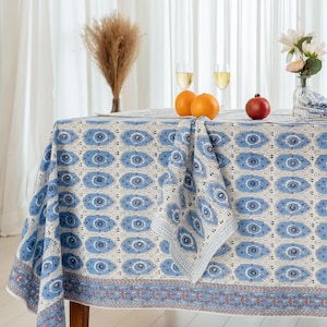 Puede incluir: Un mantel redondo con un diseño de ojo azul y blanco, con una servilleta a juego. La mesa está puesta con naranjas, una granada y copas de vino. Un jarrón con flores y hierba seca completan la escena.