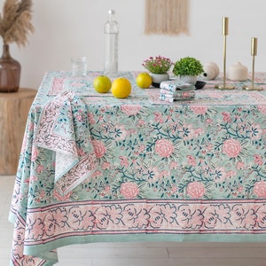 May include: A green tablecloth with a pink floral pattern and a white border. The tablecloth is laid out on a table with a glass of water, lemons, and candles.