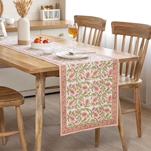 Puede incluir: Una mesa de comedor de madera con un camino de mesa con estampado floral. El camino de mesa presenta un fondo blanco con diseños florales rosas y verdes. La mesa está puesta con platos, cubiertos y una copa de vino.