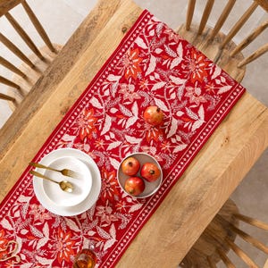 May include: A red table runner with a floral pattern in white and orange, set on a wooden table. The table is set with white plates, gold silverware, and a bowl of pomegranates. A glass of liquid is also on the table.
