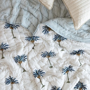 May include: A white quilted bedspread with a pattern of blue palm trees. The bedspread is paired with a blue and white striped quilt and two white pillows with a quilted design. The bedding has a tropical theme.