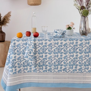 Op de afbeelding: Een blauw-wit tafelkleed met een ginkgo-bladpatroon. Het tafelkleed ligt op een tafel met fruit en een glas water.