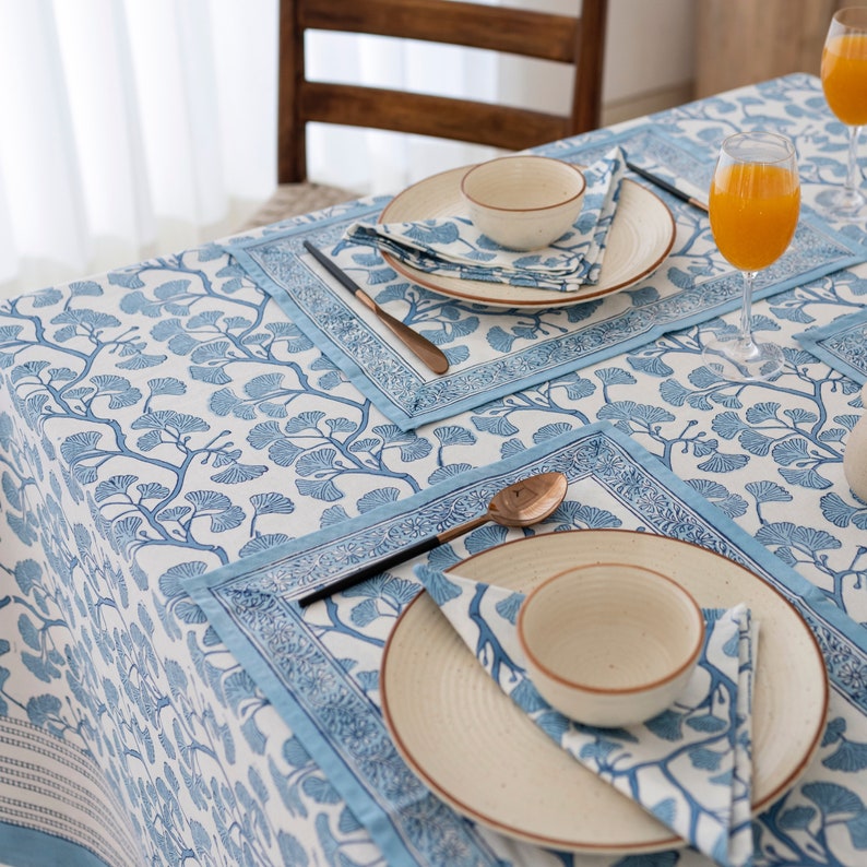 Puede incluir: Un mantel blanco con un patr&oacute;n floral azul y manteles individuales azules. Hay dos cubiertos con platos, cuencos y servilletas blancas. La mesa est&aacute; puesta para una cena informal.